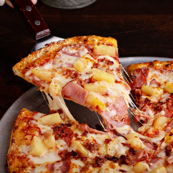 A slice of Hawaiian pizza with ham, pineapple, and melted cheese being lifted from a pan.