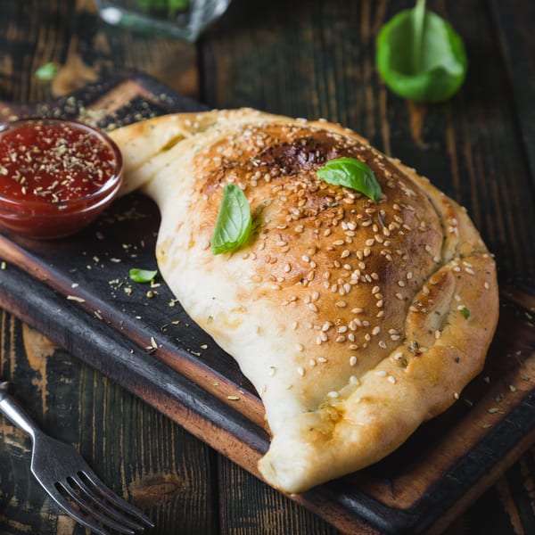 Golden-brown calzone with sesame seeds and basil, served with a side of red sauce on a wooden board.