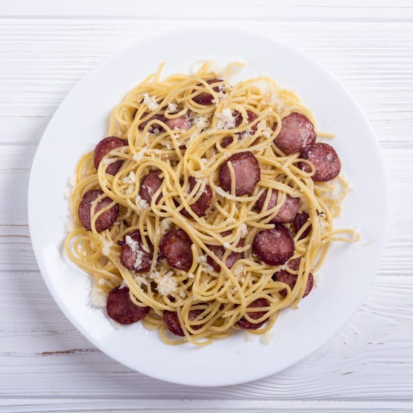 Spaghetti with sliced sausage and grated Parmesan cheese on a white plate.